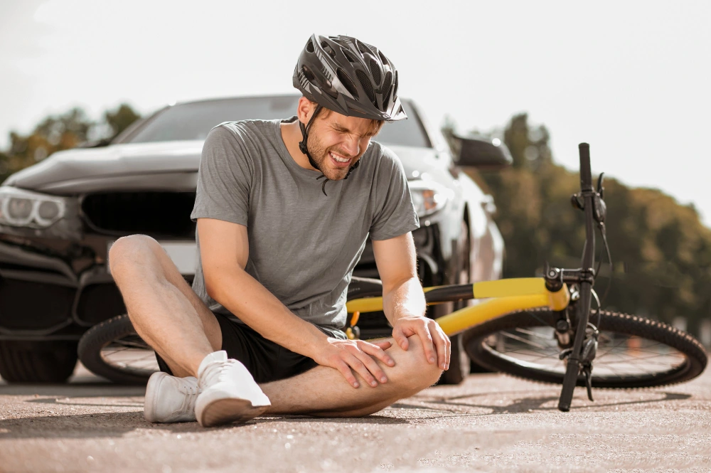 young adult injured cyclist in helmet sitting holding knee on road